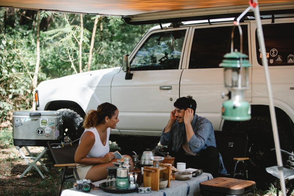 Cheerful multiracial couple communicating while sitting at table near camping van with awning in forest with green trees in sunny day