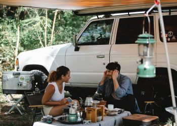 Cheerful multiracial couple communicating while sitting at table near camping van with awning in forest with green trees in sunny day