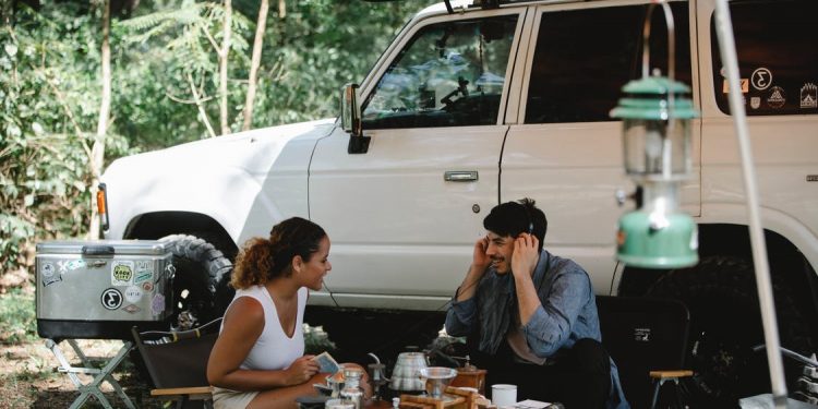 Cheerful multiracial couple communicating while sitting at table near camping van with awning in forest with green trees in sunny day