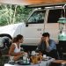 Cheerful multiracial couple communicating while sitting at table near camping van with awning in forest with green trees in sunny day